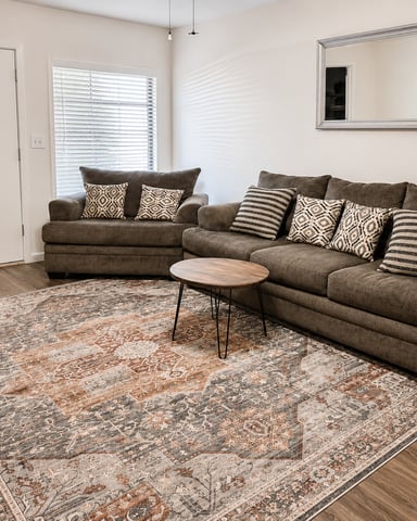 Living room with brown sectional sofa, patterned area rug, round wood coffee table, and framed mirror on white walls